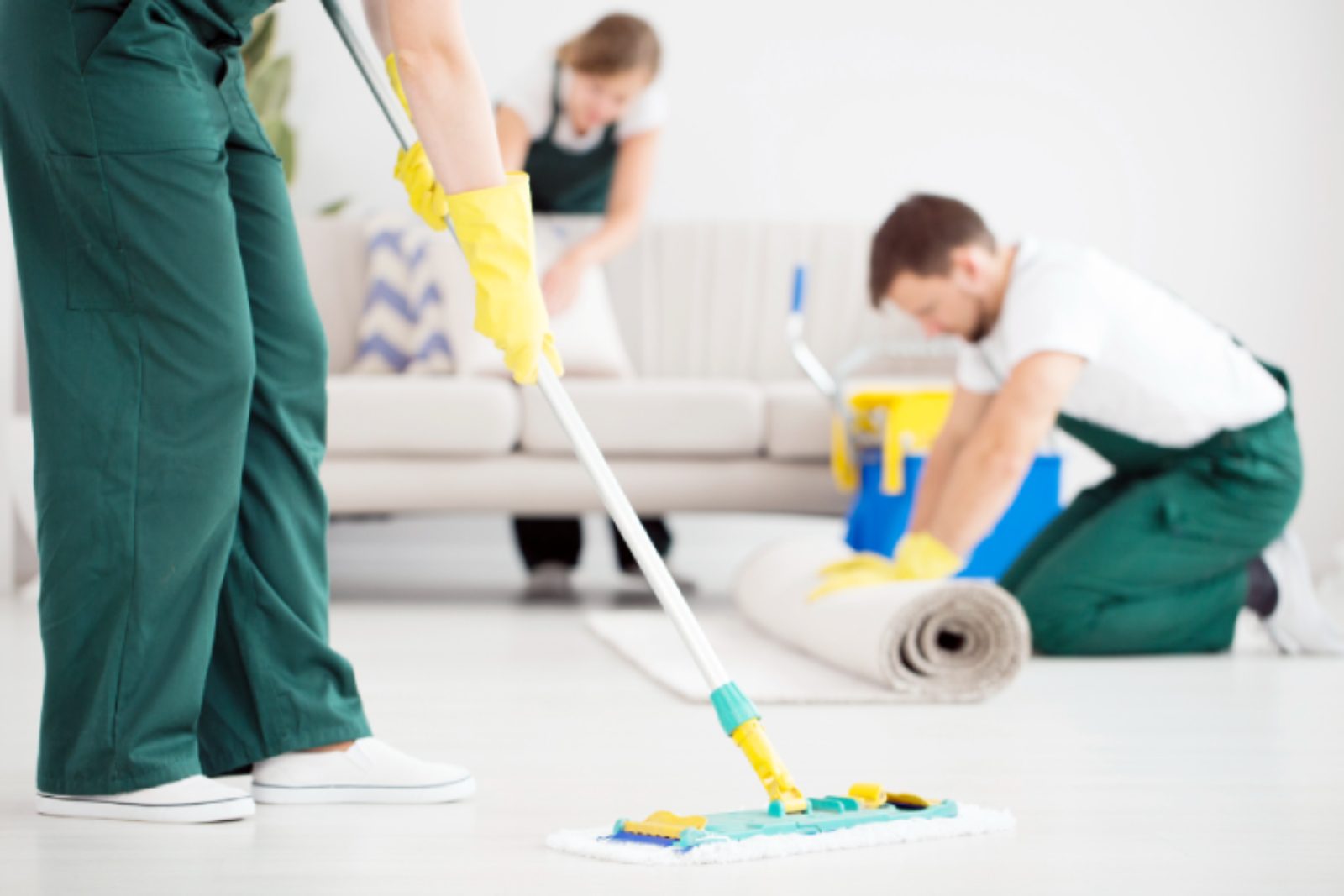Three people in green uniforms are cleaning a room; one is mopping the floor while the others organize a rolled carpet near a couch.