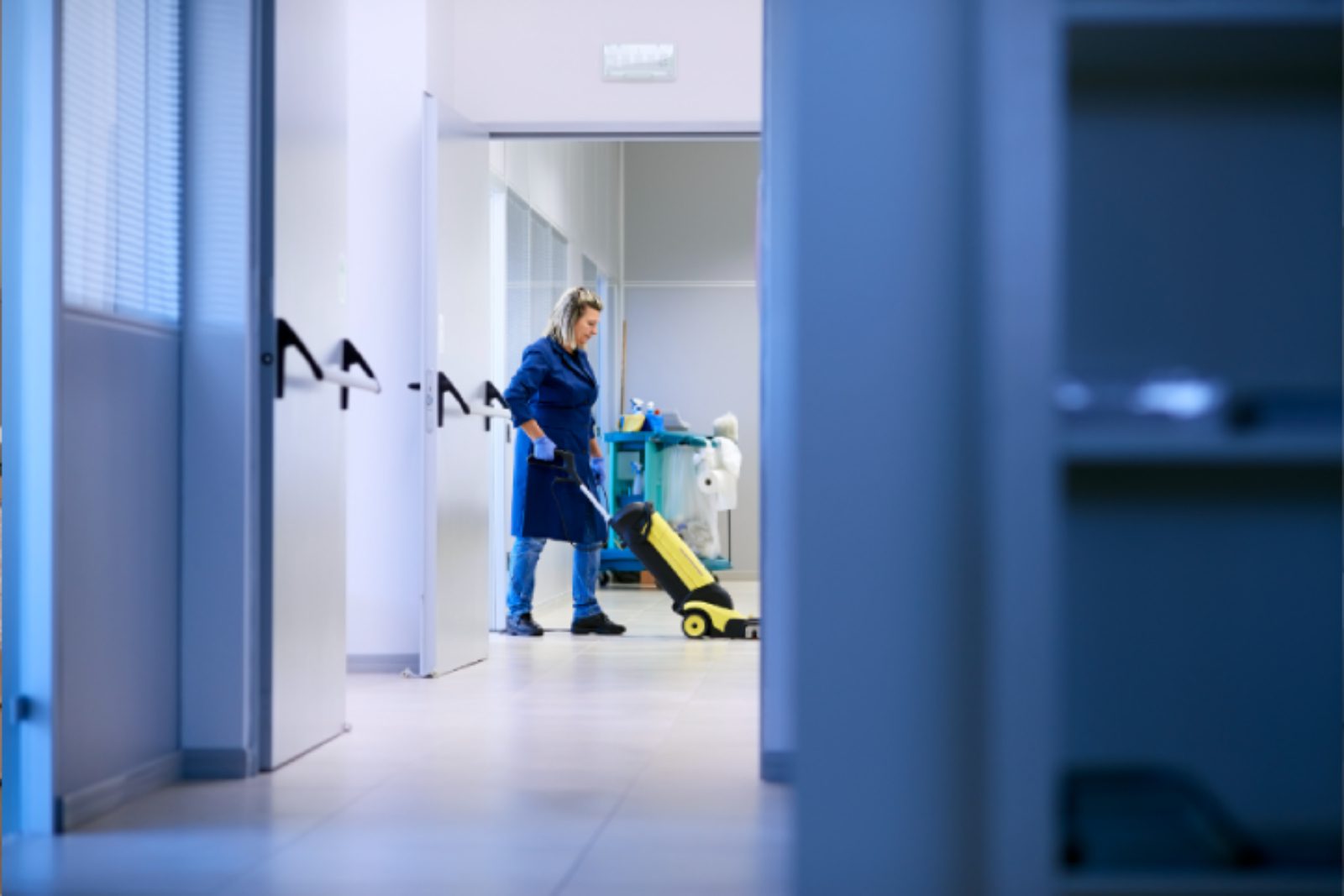 A person in blue uniform cleaning a hallway floor with a machine. A cleaning cart is visible in the background.
