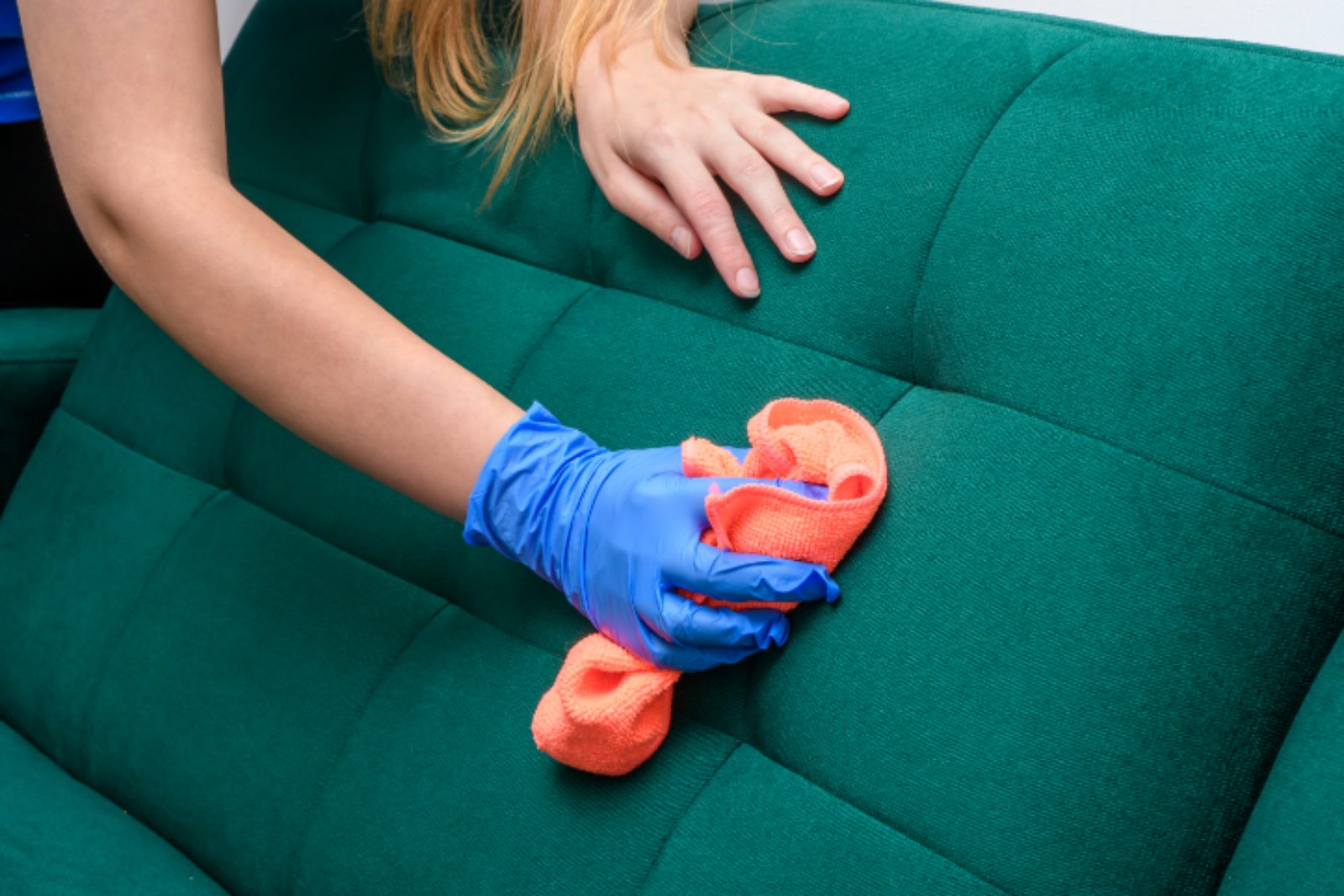 Person wearing blue gloves cleans a green couch with an orange cloth.