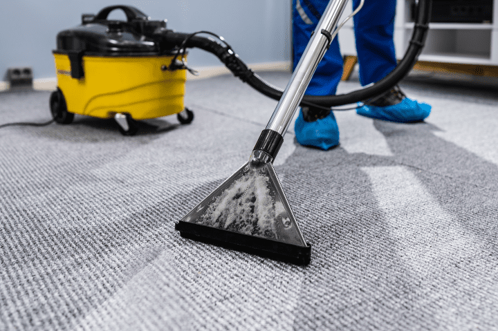 A person uses a carpet cleaning machine on a gray carpet. A yellow vacuum is in the background, and the individual wears blue shoe covers.
