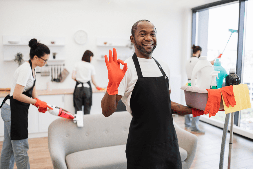 A group of four people in aprons are cleaning a room. One person in the foreground holds cleaning supplies and gives an OK gesture. Others are vacuuming and tidying the space.