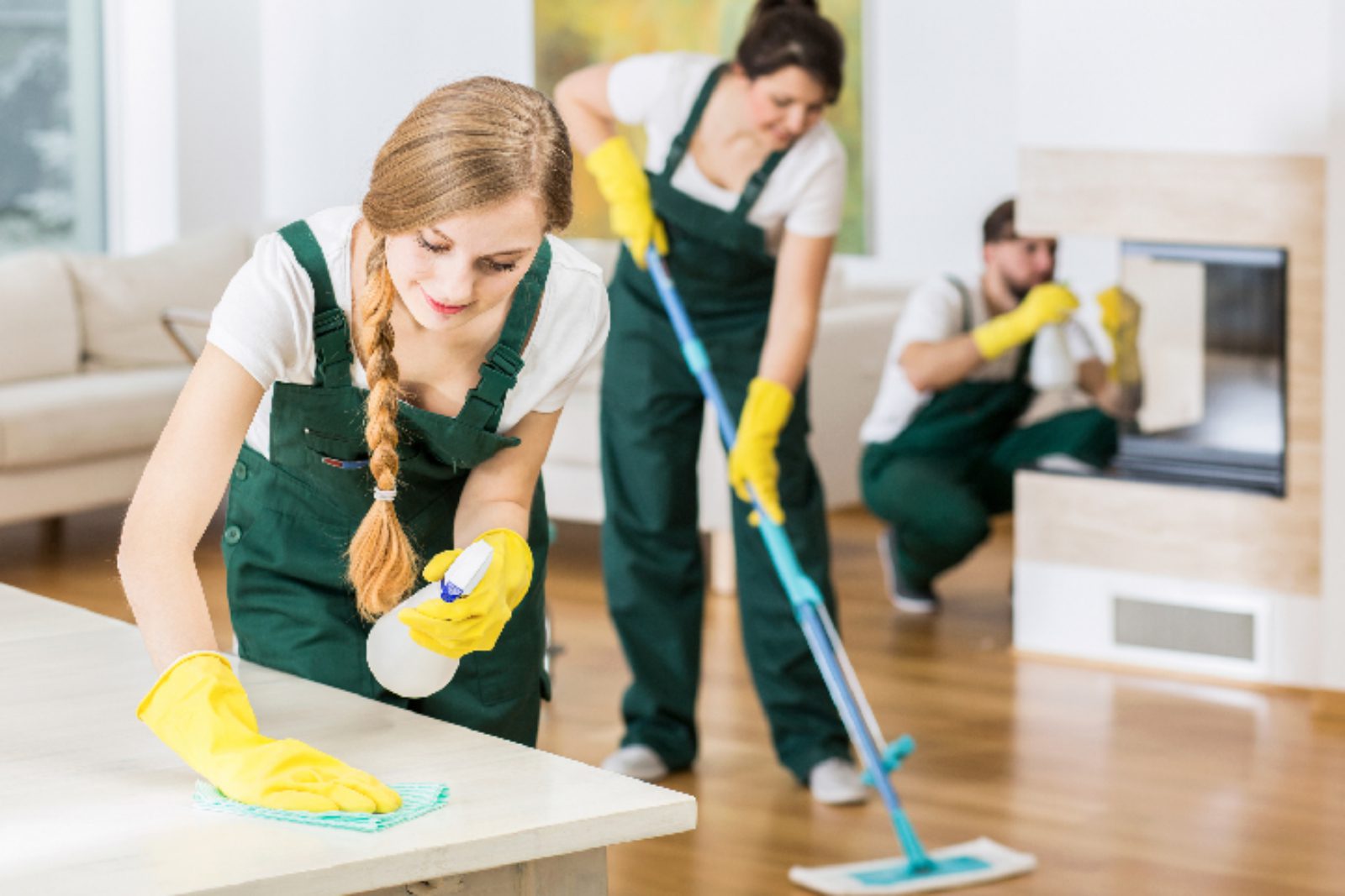Three people wearing green overalls and yellow gloves clean a room; one wipes a table, another mops the floor, and the third cleans a mirror.