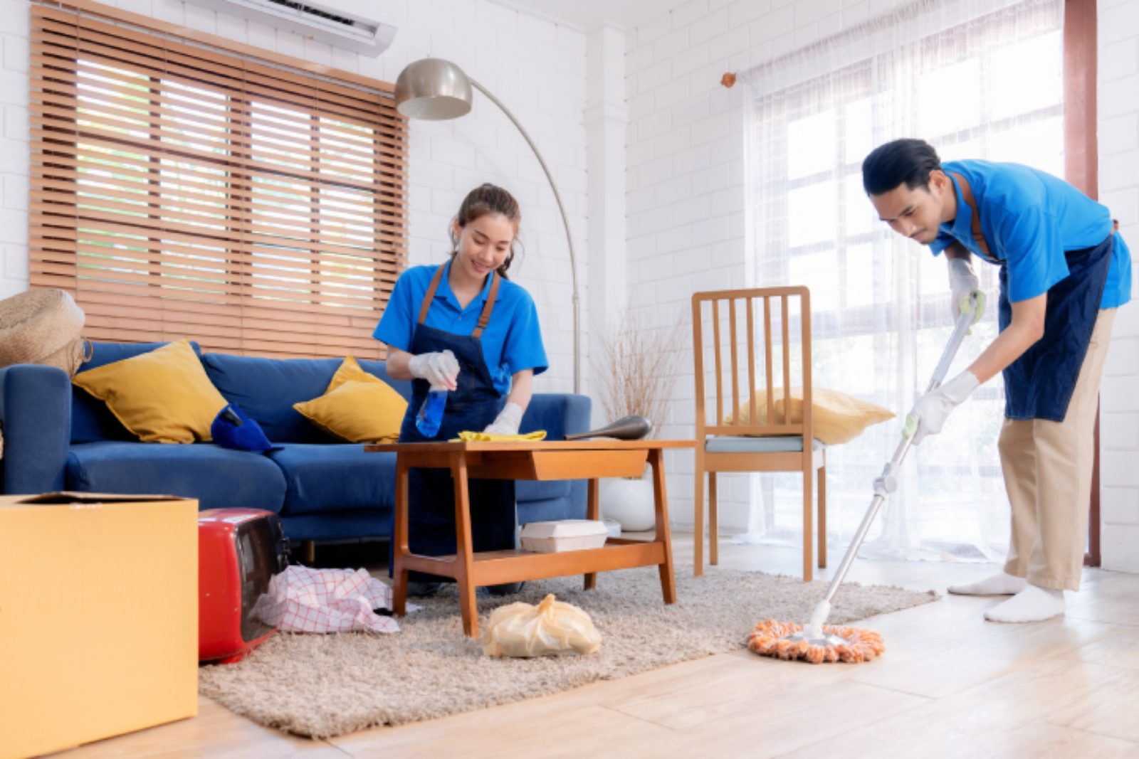Two people in blue uniforms clean a living room. One is polishing a table, while the other mops the floor. The room has a sofa, coffee table, and window with blinds.