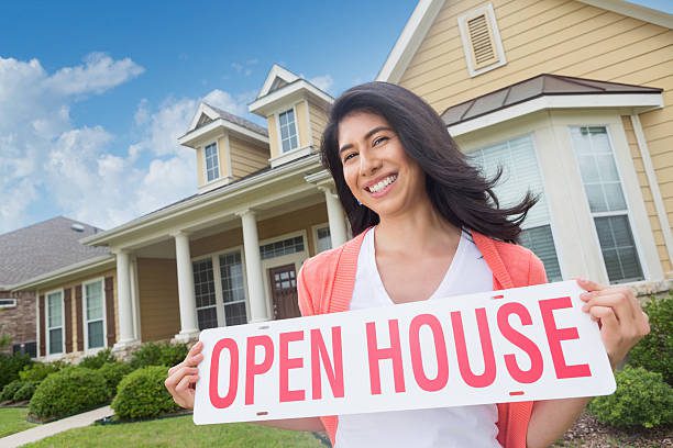 A smiling woman holding an open house sign in front of a house.