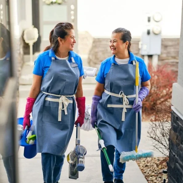 Two women in cleaning uniforms walking and smiling with cleaning tools.